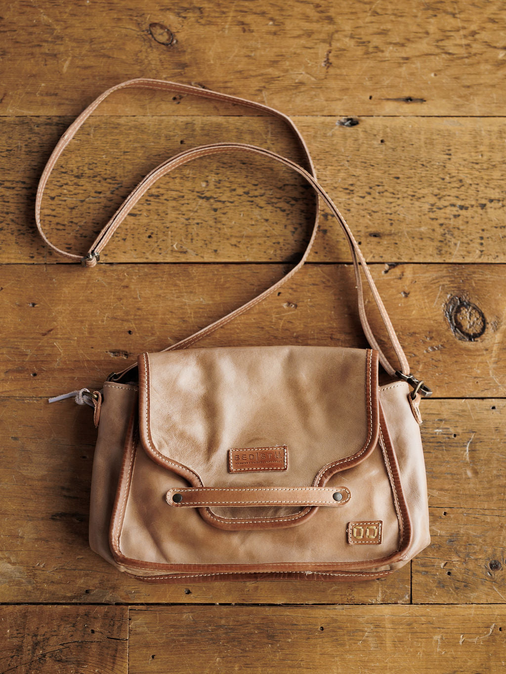 The Lomita crossbody bag by Savannah Sevens western life{&}style, crafted in rustic oat leather with a flap closure, shoulder strap, detailed stitching, and metal accents, rests on a rustic wooden surface.