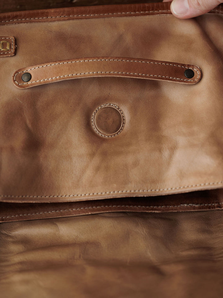Close-up of the inside flap of Savannah Sevens western life{&}style's The Lomita crossbody bag in rustic oat leather, showing visible stitching, two metal rivets, a central circular indentation, and a hand holding the edge.