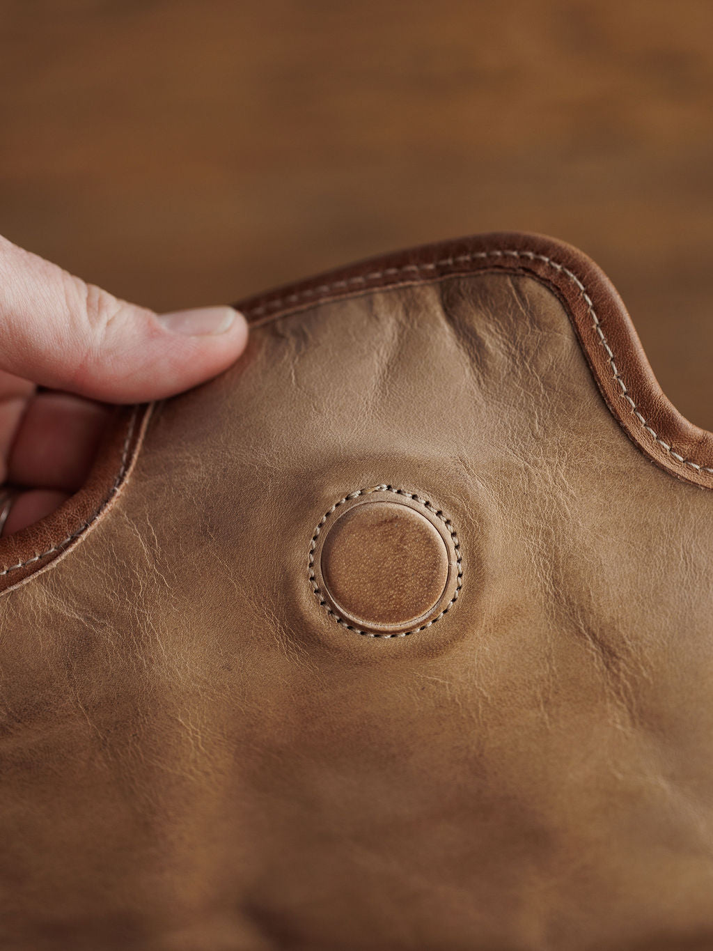 A close-up of a hand holding rustic oat leather with visible stitching and a circular detail, ideal for crafting The Lomita crossbody bag by Savannah Sevens western life{&}style. The leather has a natural, textured look.