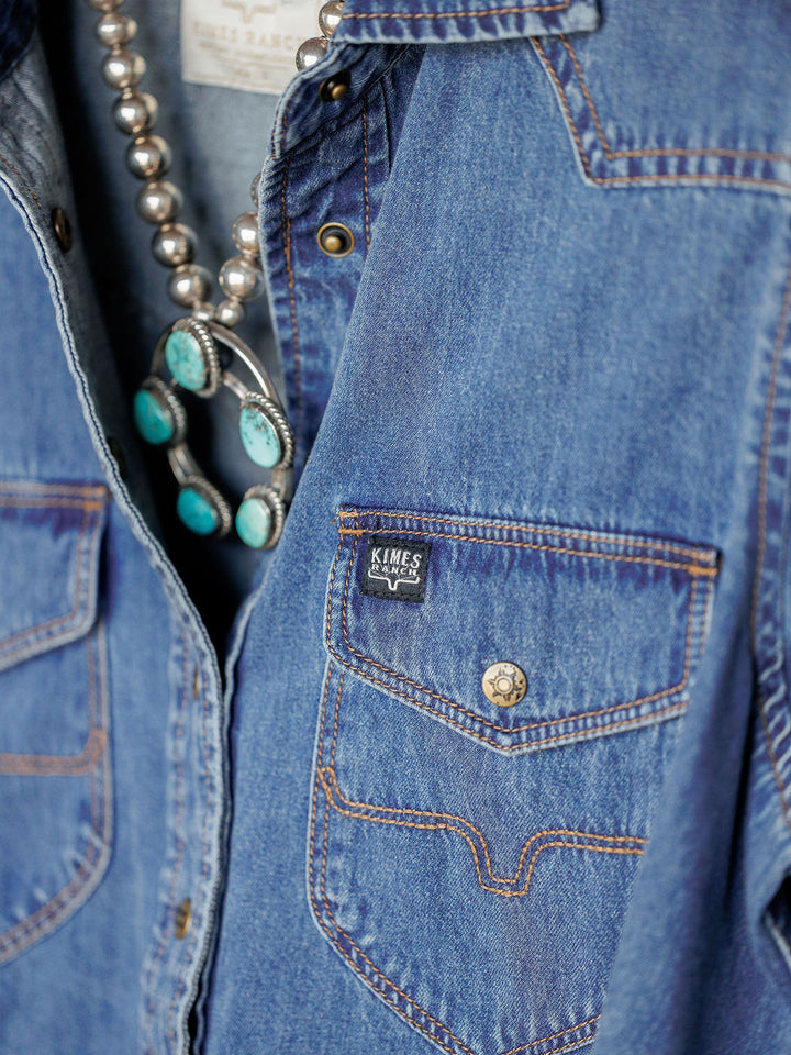 Close-up of Savannah Sevens western life{&}style's The Dalles relaxed fit denim snap button top with brown stitching, metal buttons, and a branded pocket. A silver necklace with turquoise stones accents the collar.