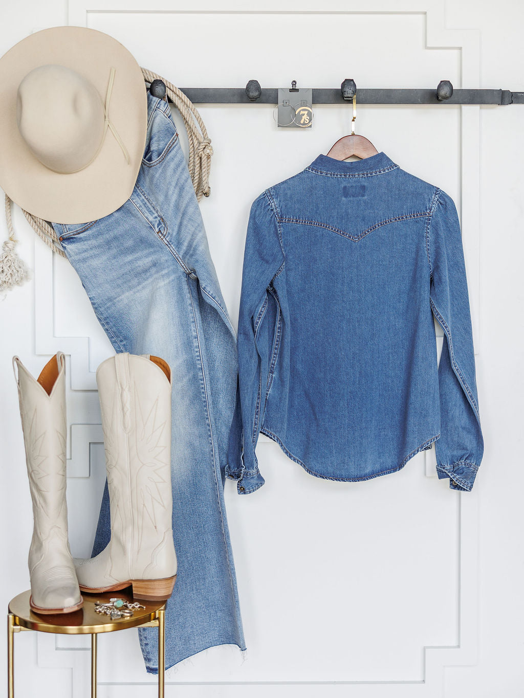 The Dalles by Savannah Sevens western life{&}style: a light beige cowboy hat, relaxed fit jeans, and a western yoke denim shirt hang above a gold stool with tall white boots, all set against a white wall with decorative molding.