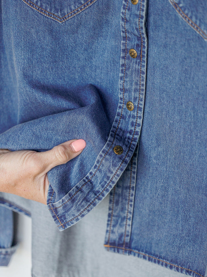 A person places their hand at the hem of The Dalles women’s relaxed fit denim shirt by Savannah Sevens western life{&}style, featuring brass buttons and visible stitching.