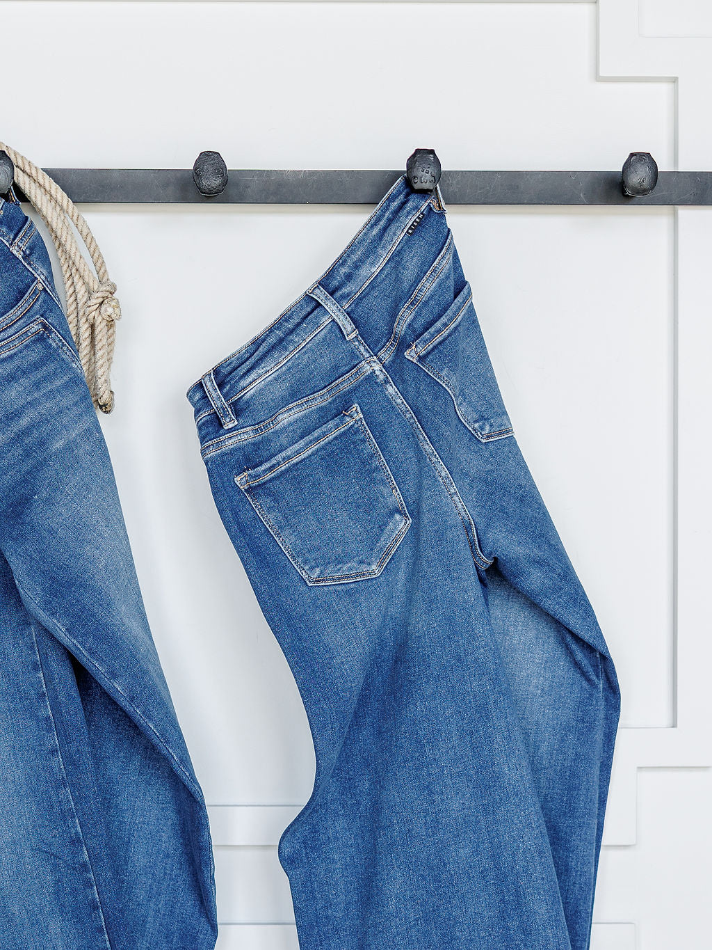 The Maren High Rise Wide Leg Jean by Savannah Sevens western life{&}style hangs on a metal rod against a white wall with decorative molding, with another pair of wide leg jeans partially visible to the left.