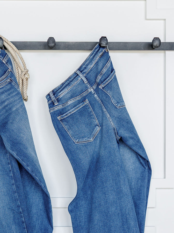 The Maren High Rise Wide Leg Jean by Savannah Sevens western life{&}style hangs on a metal rod against a white wall with decorative molding, with another pair of wide leg jeans partially visible to the left.