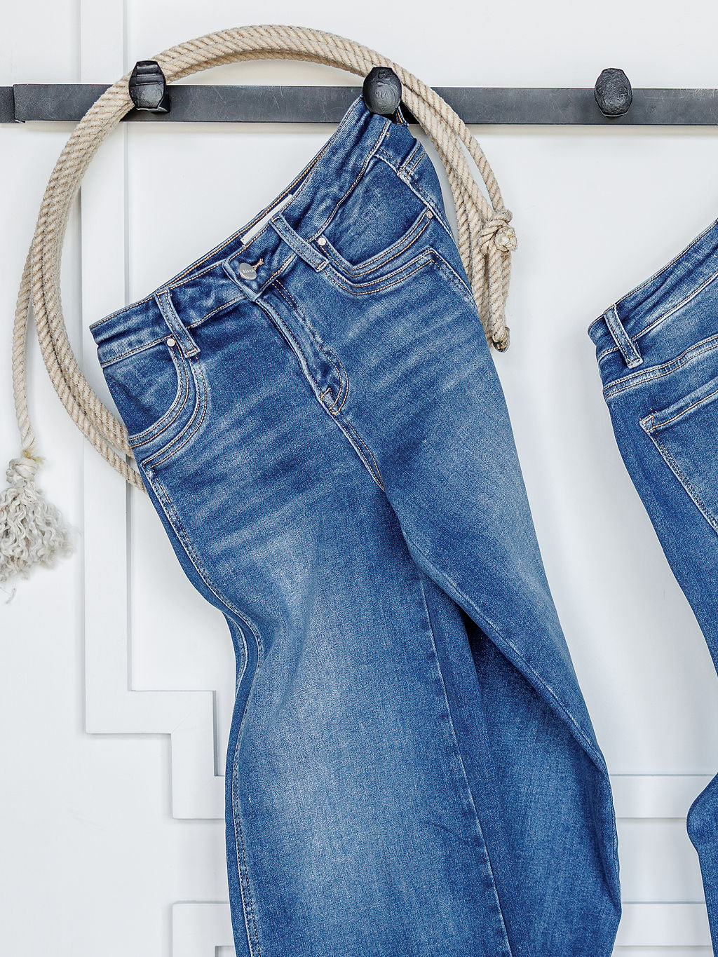 A pair of The Maren High Rise Wide Leg Jean by Savannah Sevens western life{&}style hangs from a white rope over a black wall hook, with part of another wide leg jean beside it against a white paneled wall.