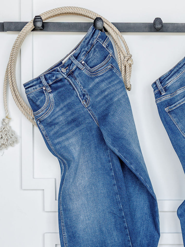 A pair of The Maren High Rise Wide Leg Jean by Savannah Sevens western life{&}style hangs from a white rope over a black wall hook, with part of another wide leg jean beside it against a white paneled wall.