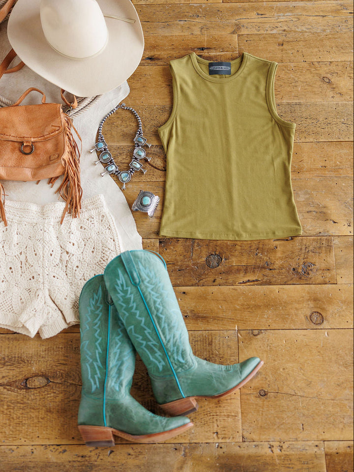 Flat lay of The Belton by Savannah Sevens western life{&}style: a country-inspired outfit featuring a beige hat, brown fringe purse, turquoise jewelry, fitted moss tank, cream crochet shorts, and teal cowboy boots on a wooden floor.