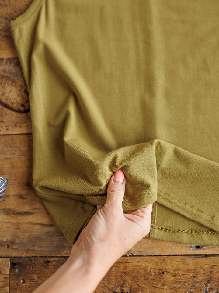 A hand gently lifts the hem of The Belton tank by Savannah Sevens western life{&}style, displayed on wood, emphasizing the texture and softness of its olive-green, stretchy fabric.