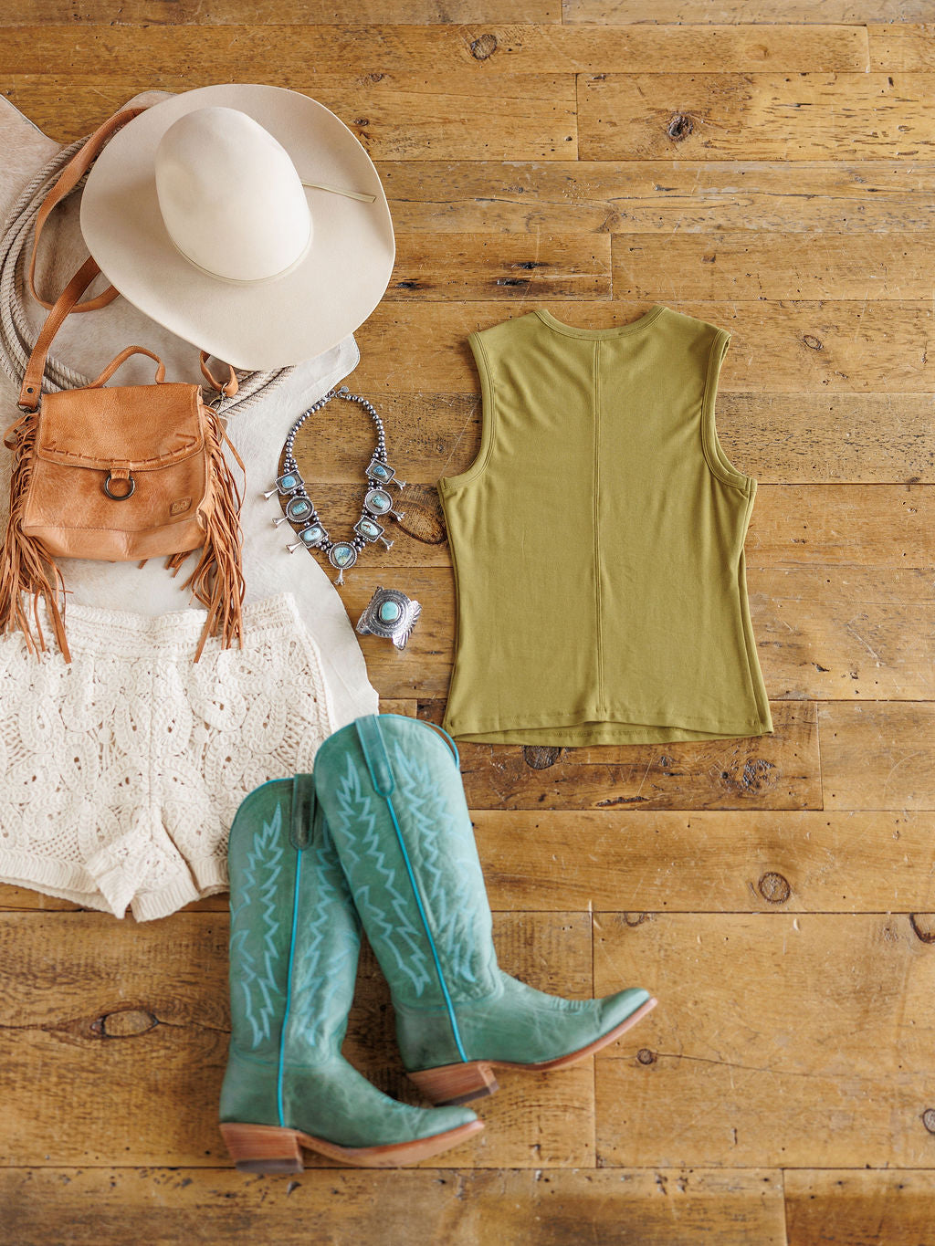 A flat lay of The Belton by Savannah Sevens western life{&}style: white crochet shorts, fitted olive-green moss tank, turquoise cowboy boots, beige wide-brim hat, tan fringe bag, and turquoise jewelry on a wooden floor.
