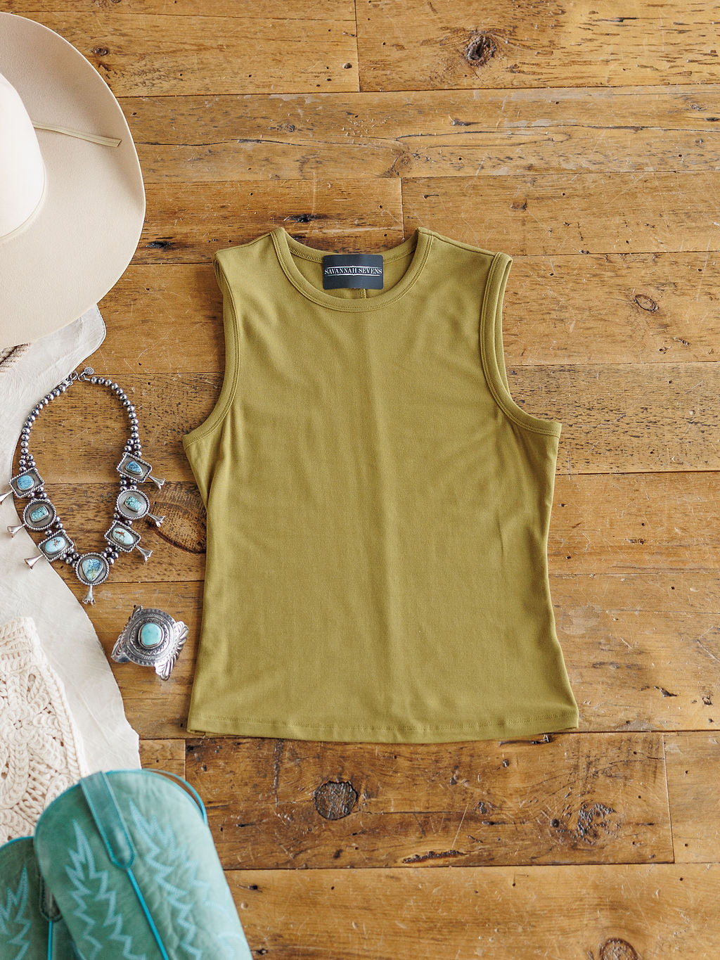 The Belton by Savannah Sevens western life{&}style is a sleeveless moss tank, shown laid flat on a wooden surface with a beige hat, turquoise jewelry, and white lace. Its stretchy fabric ensures a comfortable, flattering fit.