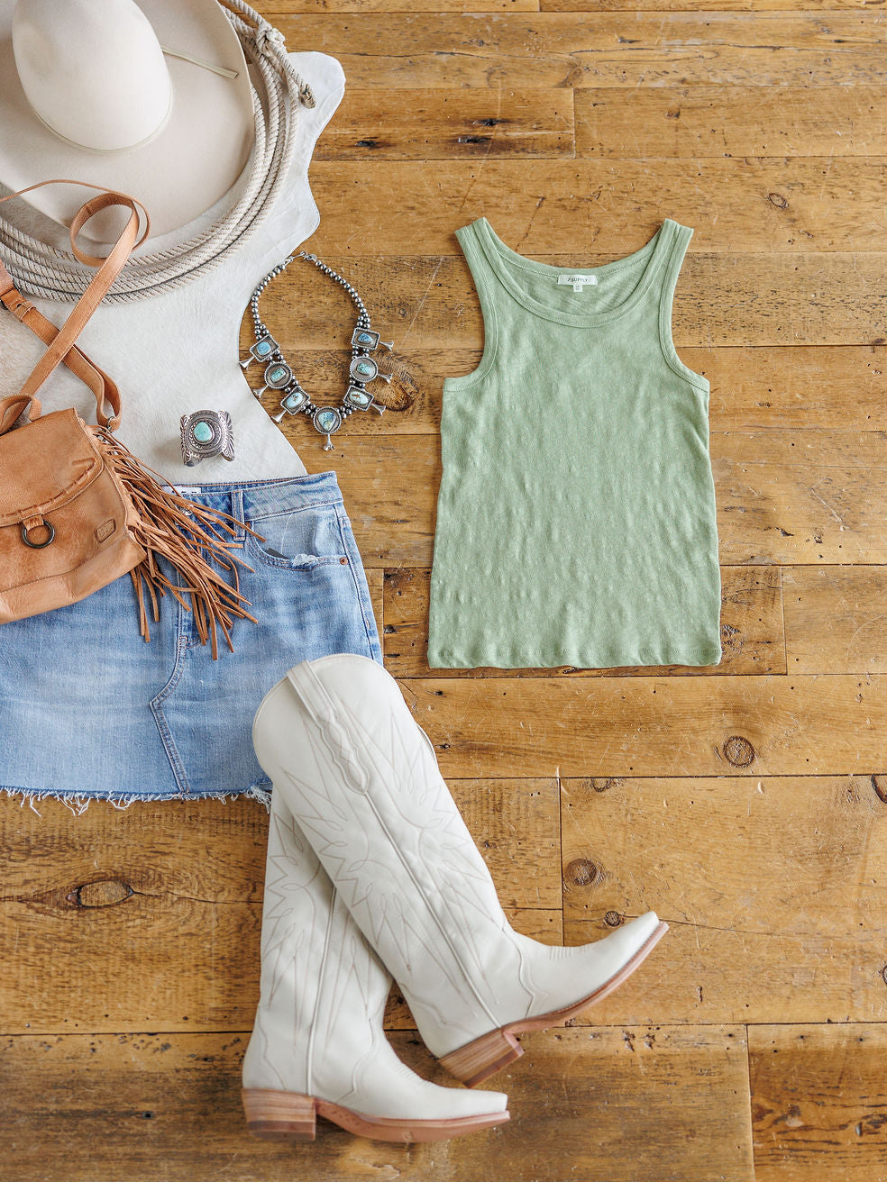 Flat lay of western-inspired outfit featuring The Asherton by Savannah Sevens western life{&}style, white cowboy boots, a light blue denim skirt, tan fringed crossbody bag, beige hat, silver jewelry, and coiled rope on a wooden floor.