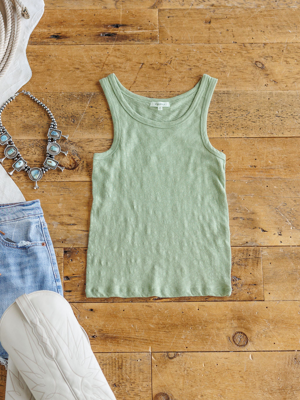 The Asherton tank by Savannah Sevens western life{&}style is styled on a wooden floor with a turquoise necklace, light blue denim shorts, and the sole of a white boot. Made from 100% cotton for all-day comfort.