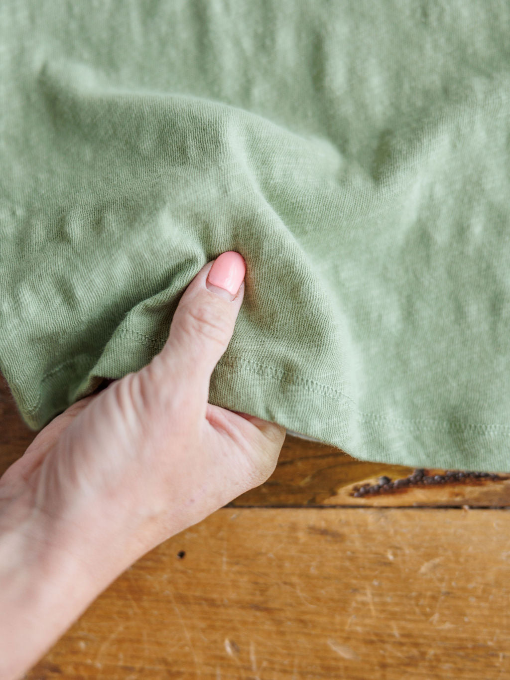 A hand pinches the hem of The Asherton tank by Savannah Sevens western life{&}style, made from 100% cotton, over a wooden surface.
