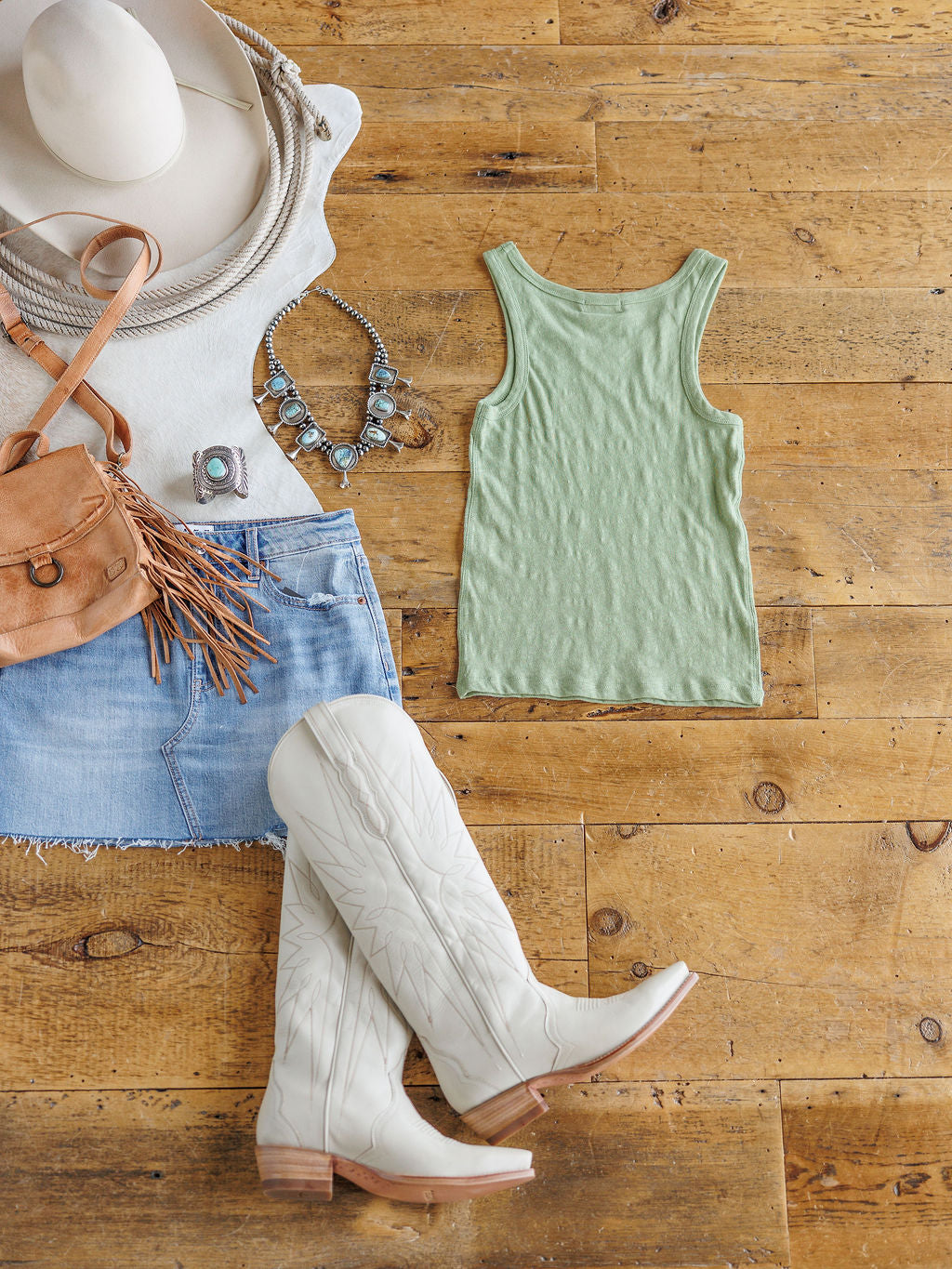 A flat lay on a wooden floor features The Asherton by Savannah Sevens western life{&}style, a sage tank in 100% cotton, paired with a denim skirt, white cowboy boots, tan fringe bag, turquoise jewelry, and a white cowboy hat.