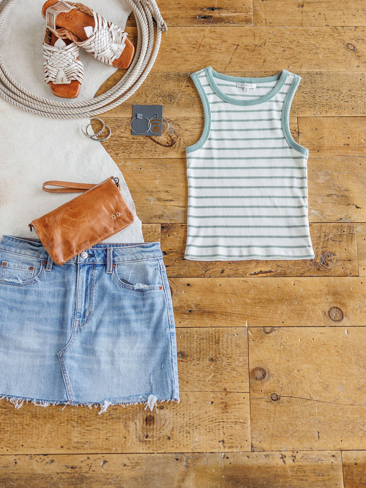 Flat lay of The Fairmont green-and-white striped ribbed tank by Savannah Sevens western life{&}style, styled with a light denim skirt, brown leather clutch, white woven sandals, silver hoops, sunglasses, and coiled rope on a wooden floor.