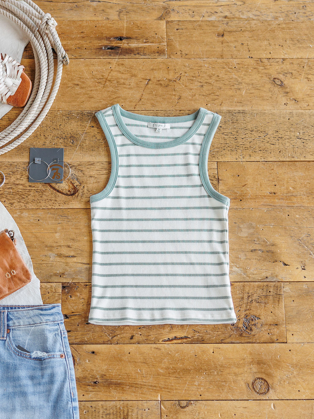 The Fairmont striped ribbed tank by Savannah Sevens western life{&}style, featuring light green and white stripes, is displayed flat on a wooden floor with blue jeans, a brown purse, and silver hoop earrings around it.