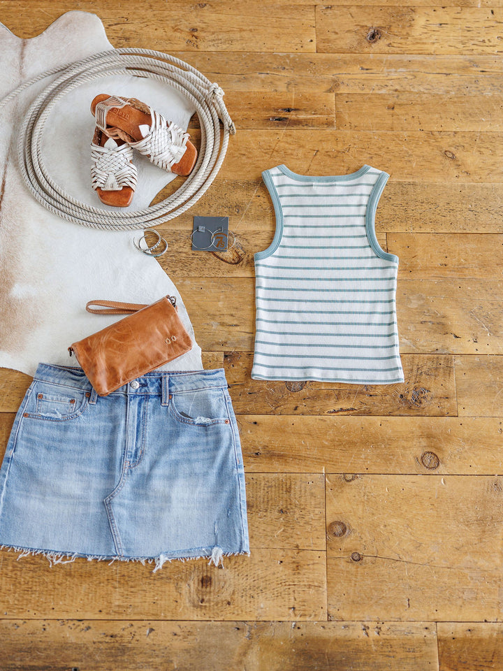 A flat lay featuring The Fairmont striped ribbed tank by Savannah Sevens western life{&}style, paired with a light blue raw-hem denim skirt, white woven sandals, tan clutch, silver jewelry, and a coiled rope on wooden flooring.