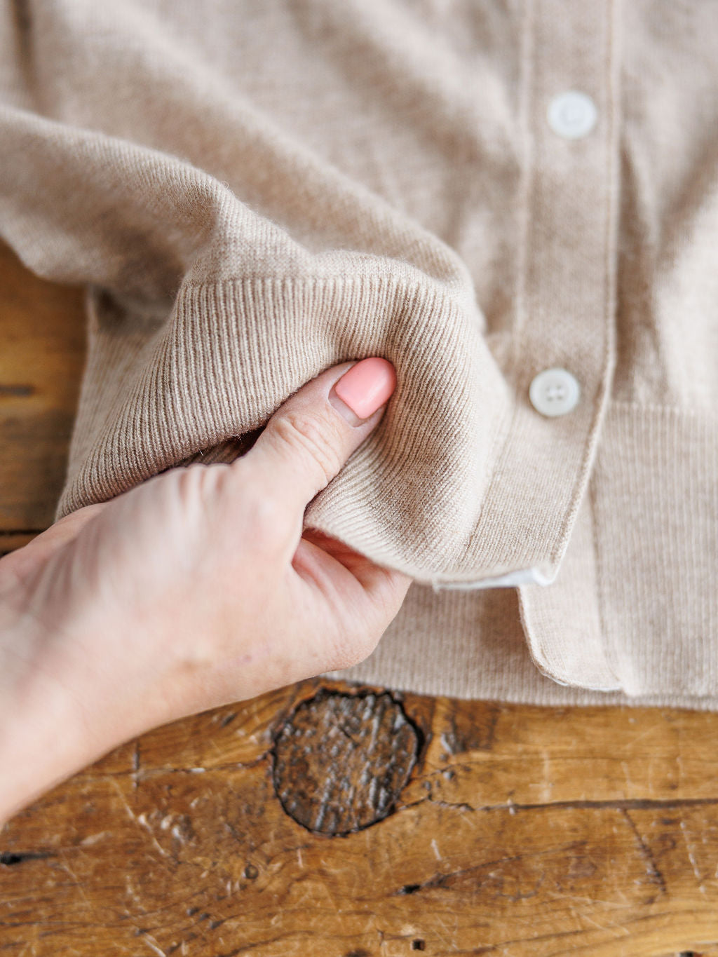 A hand  gently pinches the lightweight beige fabric of The Moran sleeveless button-up top by Savannah Sevens western life{&}style, displayed on a rustic wooden surface.