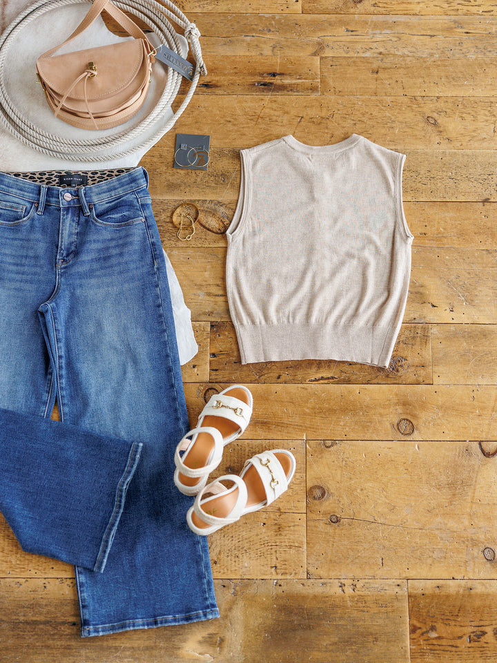 Flat lay on wood floor featuring The Moran by Savannah Sevens western life{&}style: blue jeans, sleeveless button-up top, white sandals with gold accents, beige crossbody bag, round hat, sunglasses, hoop earrings & bracelet.