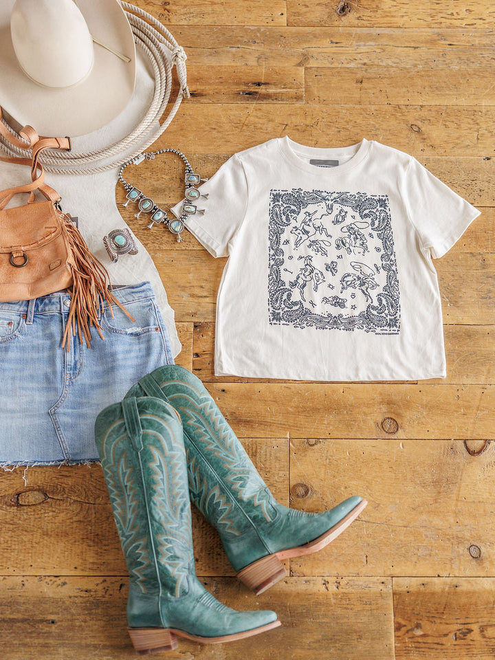 Flat lay of a casual Western outfit featuring Savannah Sevens western life{&}style's The Paisley Cowboy tee, white hat, tan fringe bag, turquoise necklace and ring, denim mini skirt, and teal cowboy boots on a wooden floor.