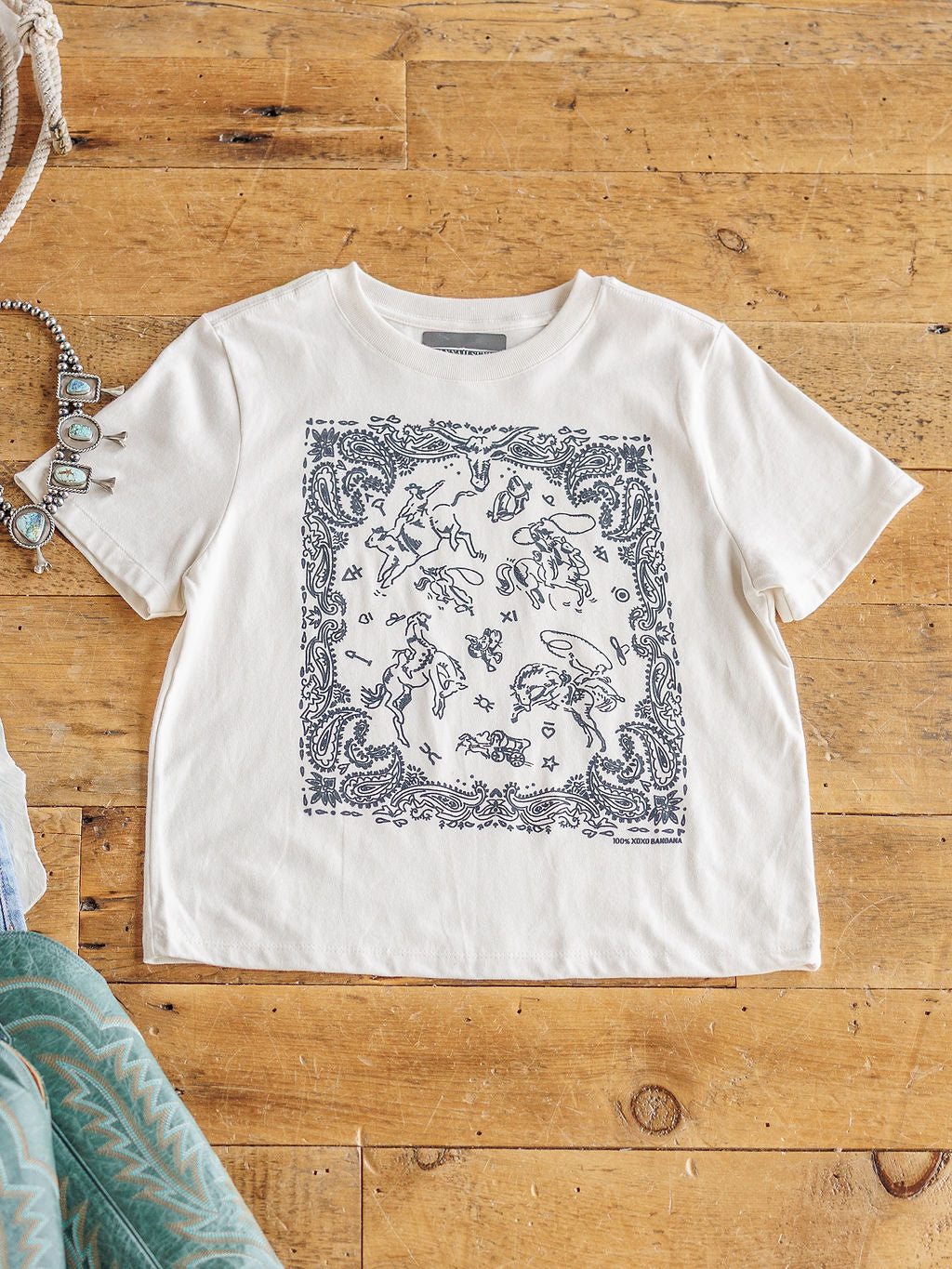 The Paisley Cowboy tee by Savannah Sevens western life{&}style is a white 100% cotton shirt with a black bandana-style print of celestial and animal graphics, shown flat on a wooden floor with jewelry and fabric nearby.