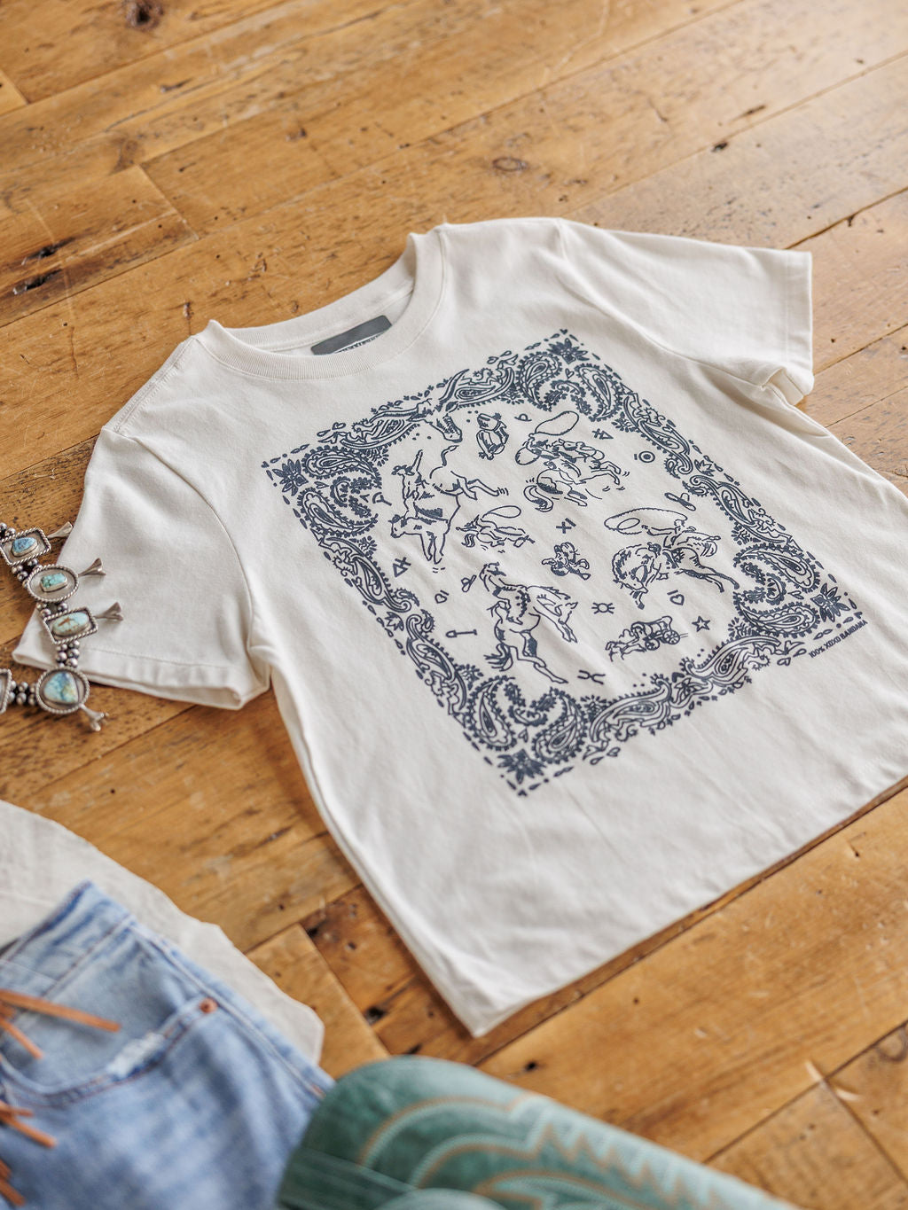 The Paisley Cowboy t-shirt by Savannah Sevens western life{&}style, made of white 100% cotton with a detailed blue graphic, is displayed on a wooden floor beside a silver bracelet and part of a pair of jeans.