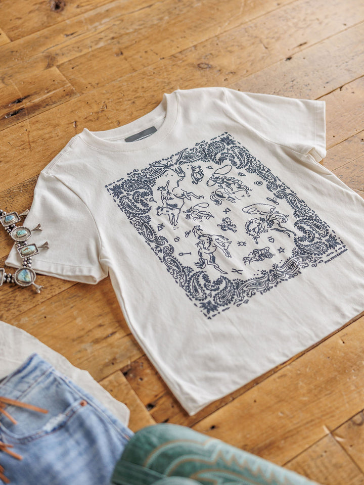The Paisley Cowboy t-shirt by Savannah Sevens western life{&}style, made of white 100% cotton with a detailed blue graphic, is displayed on a wooden floor beside a silver bracelet and part of a pair of jeans.