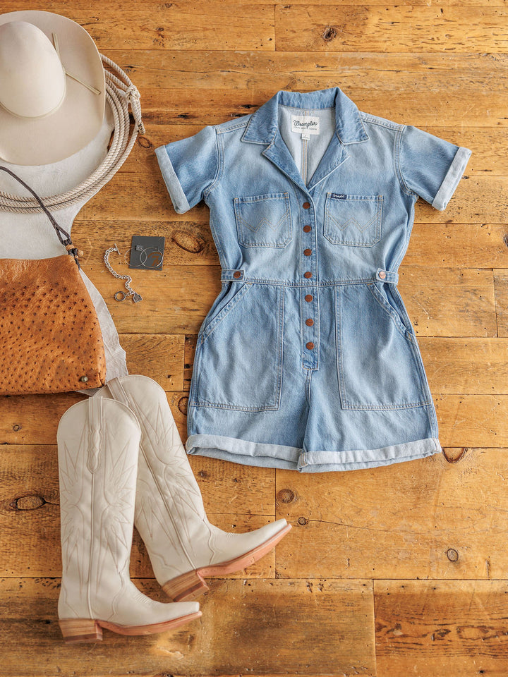 Flat lay of The Kennard medium light wash short sleeve denim romper by Savannah Sevens western life{&}style, styled with cream cowboy boots, a tan leather purse, silver jewelry, a white cowboy hat, and coiled rope on a wooden floor.