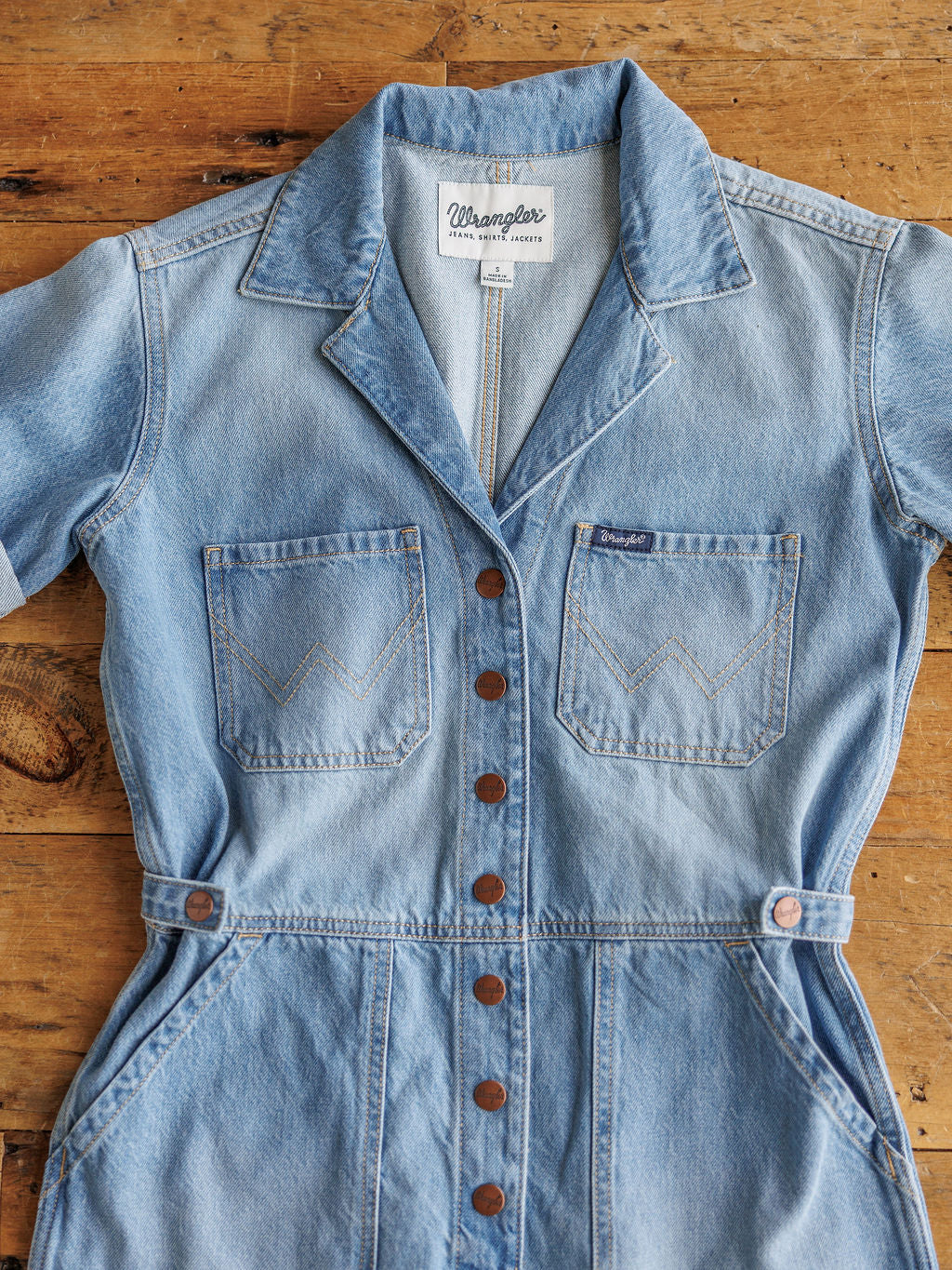 The Kennard by Savannah Sevens western life{&}style: a medium light wash, short sleeve denim romper with brown buttons, chest pockets, and a collar, shown laid flat on a wooden surface.