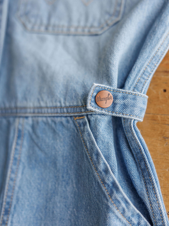 Close-up of The Kennard by Savannah Sevens western life{&}style: medium light wash denim fabric with a metal button at the belt loop, hinting at romper details. A wooden background is partially visible.