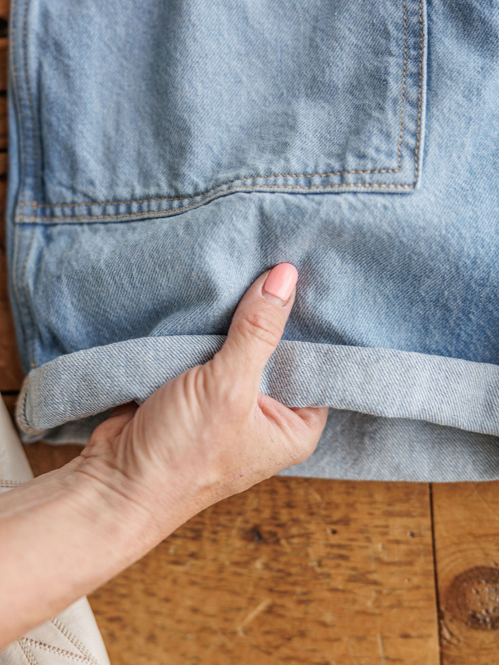 A hand  holds the cuff of The Kennard medium light wash jeans by Savannah Sevens western life{&}style, displaying the folded hem on a wooden surface.