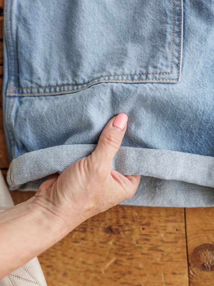 A hand  holds the cuff of The Kennard medium light wash jeans by Savannah Sevens western life{&}style, displaying the folded hem on a wooden surface.