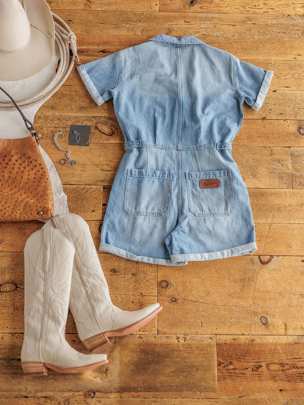 The Kennard by Savannah Sevens western life{&}style, a short-sleeve denim romper in medium light wash, is laid flat on a wooden floor with white cowboy boots, a brown leather purse, silver jewelry, and a white cowboy hat.
