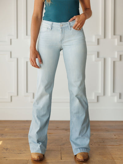 A woman wears Savannah Sevens western life{&}style's The Dani Perfect Rise Trouser Jean *Exclusive* in light wash, a teal sleeveless top, and tan shoes while standing on a wooden floor with her face not visible.