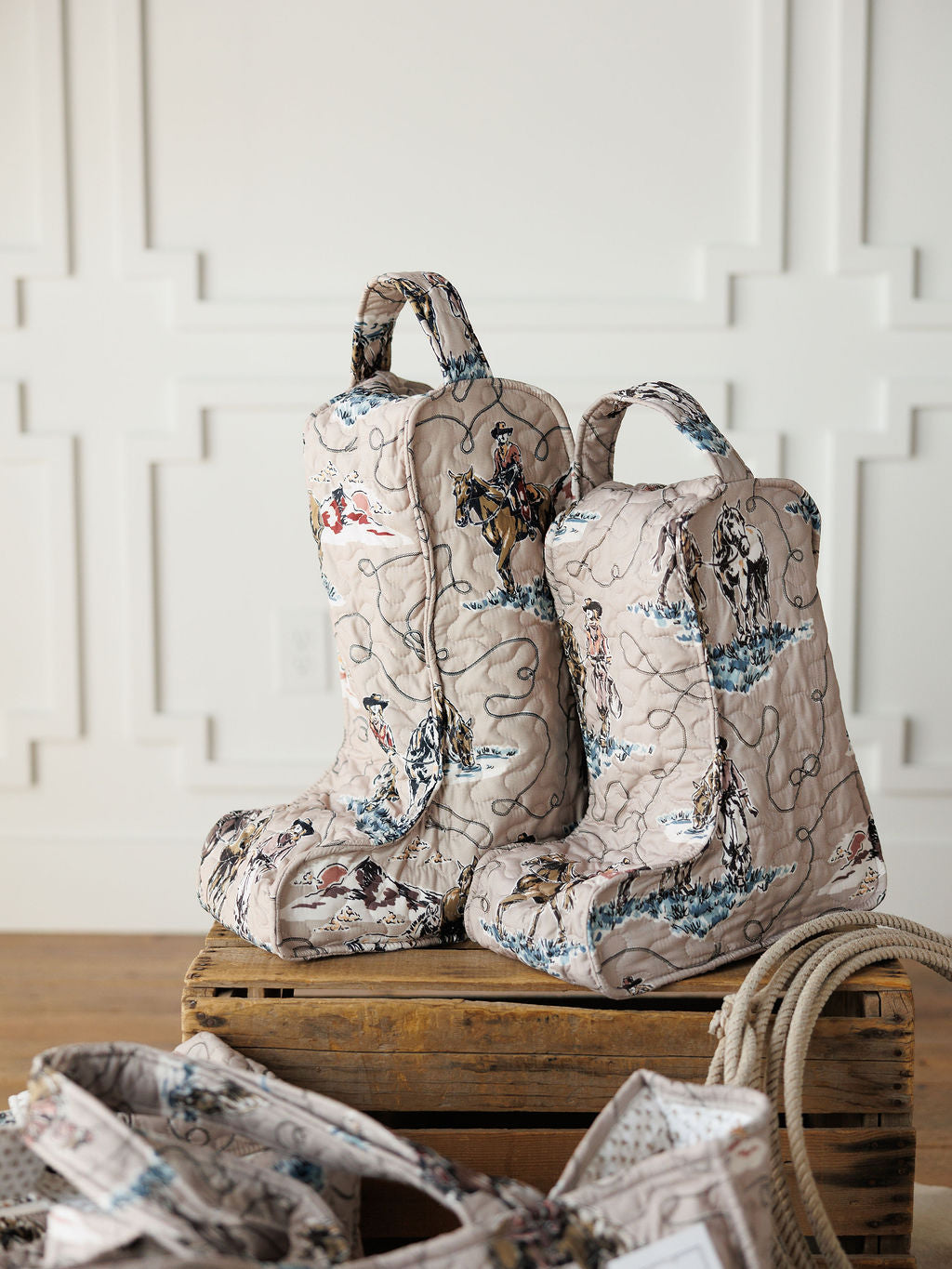 Two quilted boot bags from the Montana Summer Collection by Savannah Sevens western life&style, with Western-themed prints and large handles, sit on a wooden crate in a bright room. Nearby are a coiled rope and matching bag.