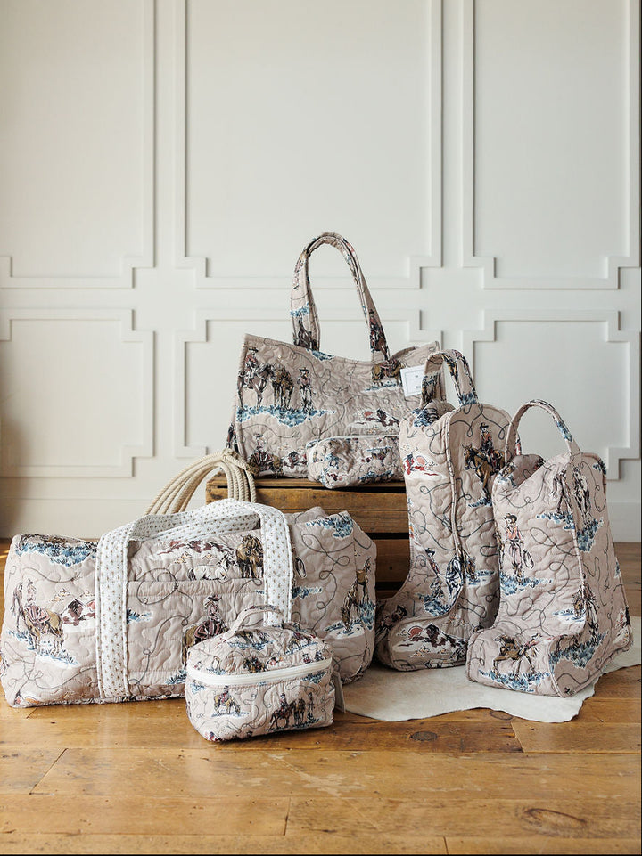 Five quilted travel bags and boots with a vintage map print from the Montana Summer Collection by Savannah Sevens western life&style are displayed on a wooden floor in front of a white paneled wall.
