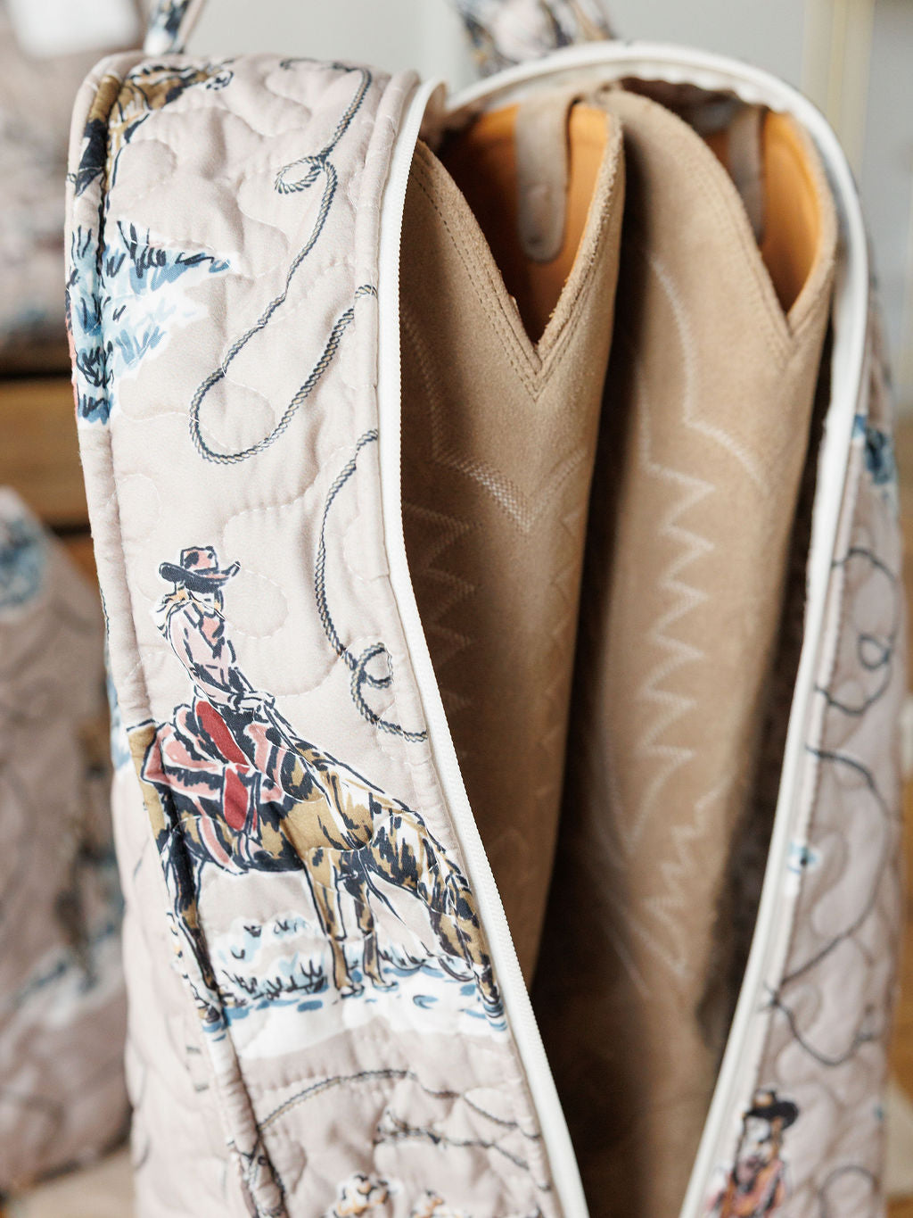 A close-up of a handmade quilt bag from the Montana Summer Collection by Savannah Sevens western life&style, featuring cowboy and horse illustrations, rope designs, and two brown zigzag-stitched padded dividers inside.