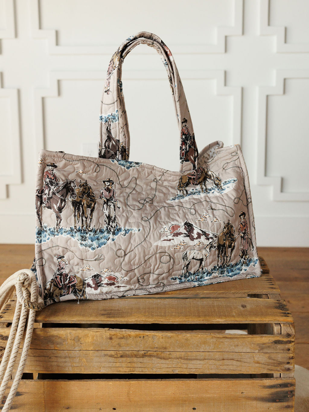 The Montana Summer Collection by Savannah Sevens western life&style features a handmade quilted bag set with cowboy and horse prints, shown on a crate with rope against a white paneled wall, highlighting its unique western style.