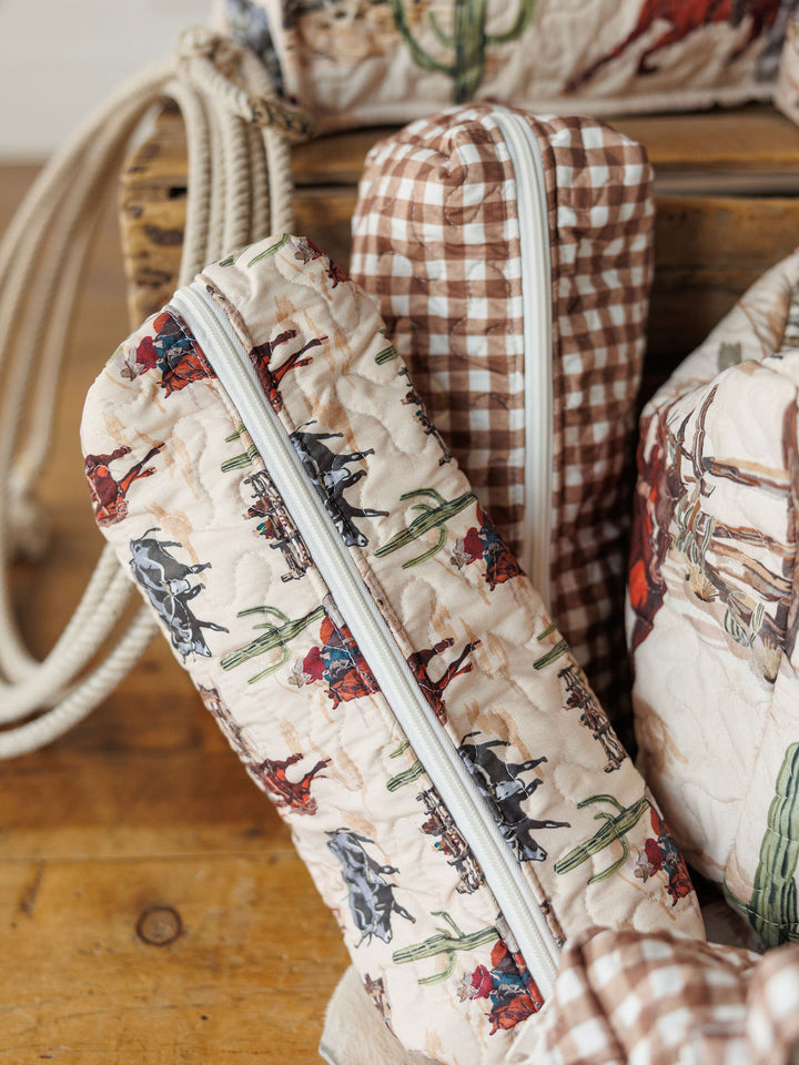 The Cowgirl Gingham Collection by Savannah Sevens western life{&}style features quilted travel bags—one with a cowboy and horse amid cacti, another in vintage gingham. Both have white zippers and are displayed on a wooden surface.