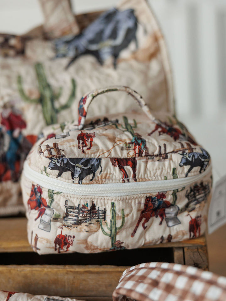 A small quilted travel bag from the Cowgirl Gingham Collection by Savannah Sevens western life&style, featuring a cowboy and horse print with cacti and ranch scenes, rests on wood beside other western-themed bags.