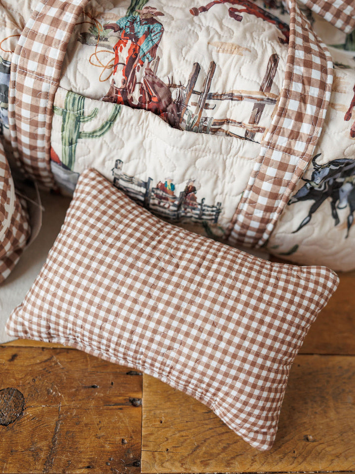 A small gingham pillow from the Cowgirl Gingham Collection by Savannah Sevens western life{&}style sits on a wooden floor beside a cowboy-themed quilt—perfect with our western bag collection.