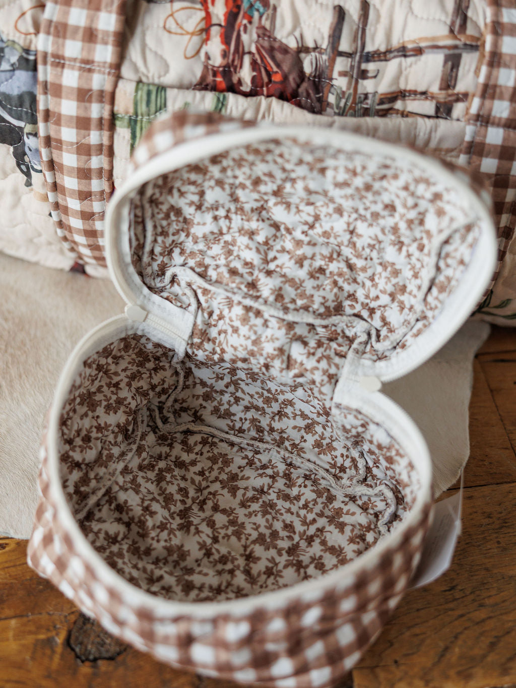 Open, empty fabric pouch with brown and white floral interior and vintage gingham exterior from Savannah Sevens western life{&}style’s Cowgirl Gingham Collection, displayed on wood beside quilted travel bags featuring horse patterns.