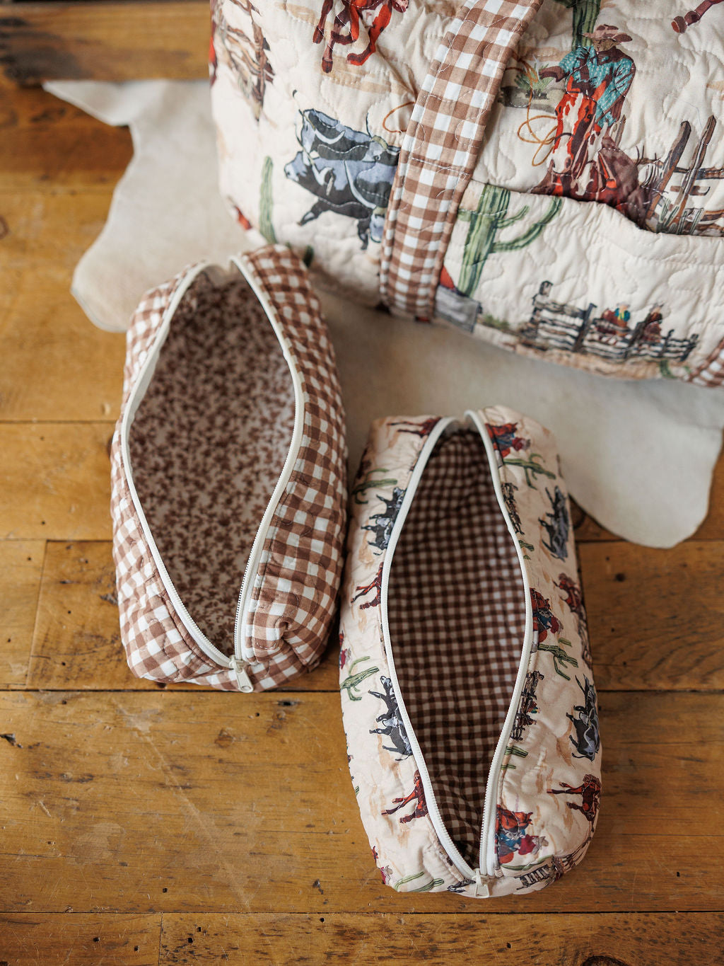 Two zippered fabric toiletry bags from the Cowgirl Gingham Collection by Savannah Sevens western life&style rest on wood—one in vintage gingham, one with a cowboy and cactus print, both with checked lining. A matching quilted travel bag is also seen.