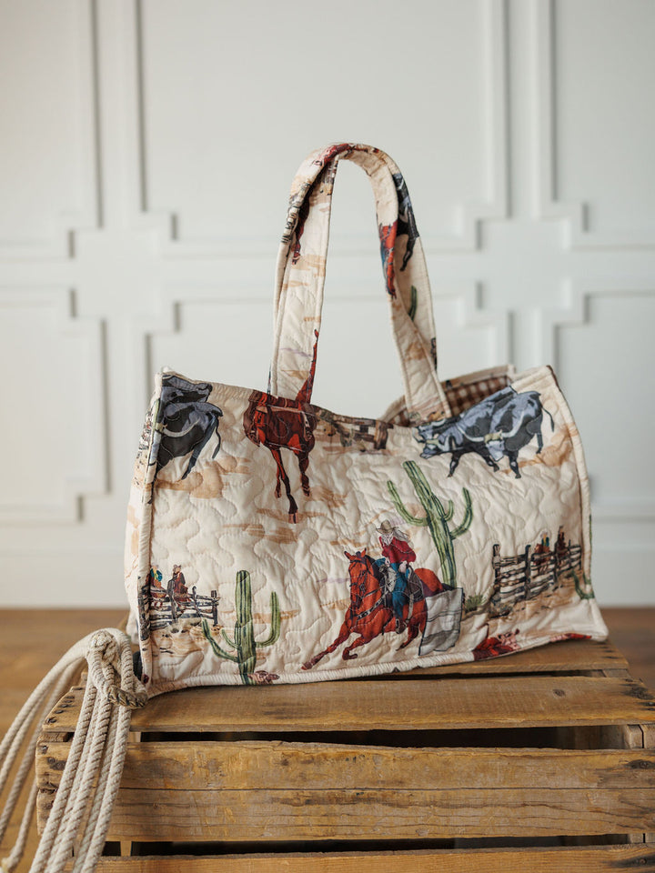 The Cowgirl Gingham Collection quilted tote by Savannah Sevens western life{&}style, adorned with cowboys, horses, cacti, and fences, is displayed on a wooden crate with a coiled rope against a white paneled wall.