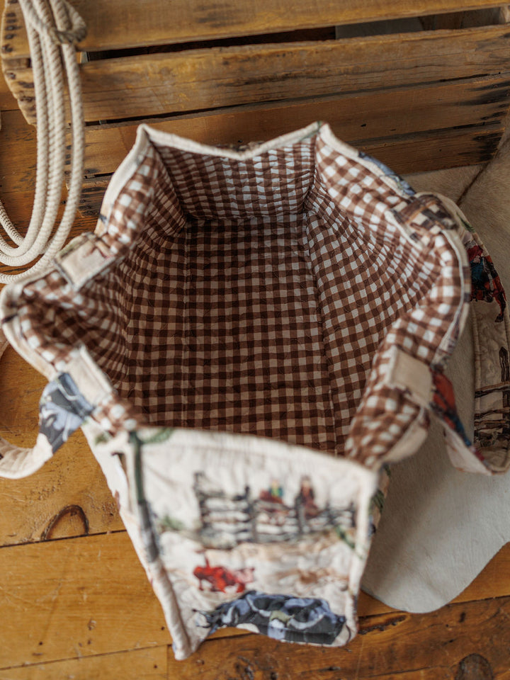 The Cowgirl Gingham tote from Savannah Sevens western life{&}style features a brown and white vintage gingham lining and a rustic, illustrated exterior pattern, pictured on wood beside rope and a light hat.