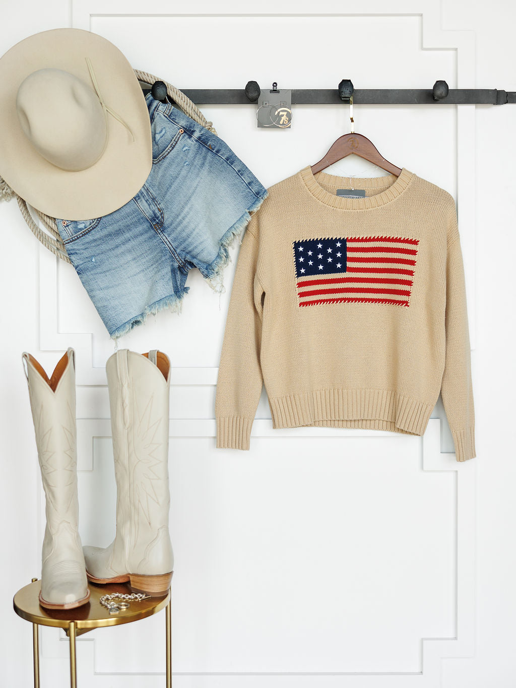 A beige cowboy hat, denim shorts, and The Original {Exclusive} embroidered American flag sweater by Savannah Sevens western life{&}style hang on a wall. Below, a gold table displays tall cream cowboy boots and jewelry.