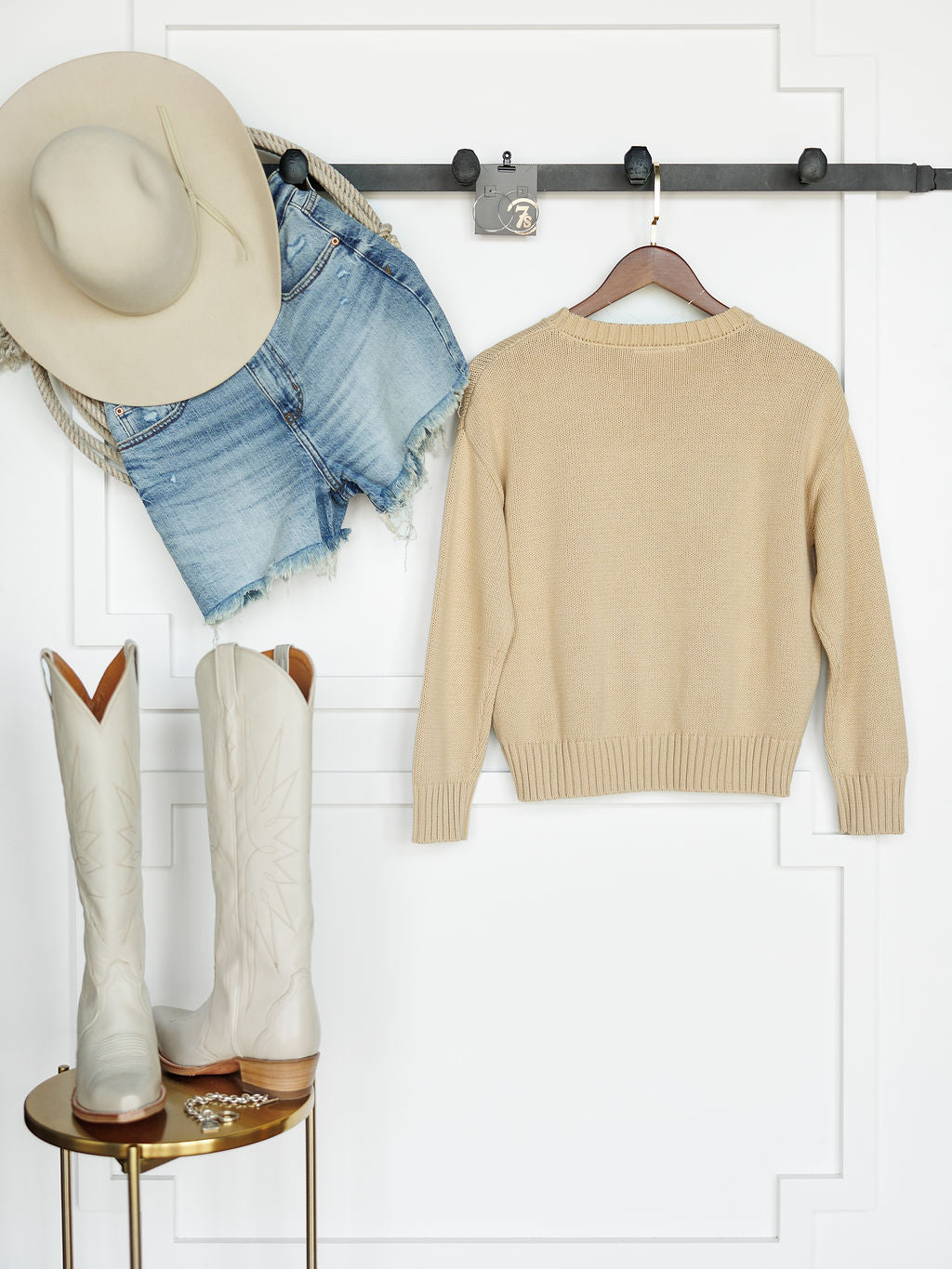 The Original {Exclusive} by Savannah Sevens western life{&}style hangs on a wall hook with cowboy boots and gold jewelry below, all set against a white paneled wall.