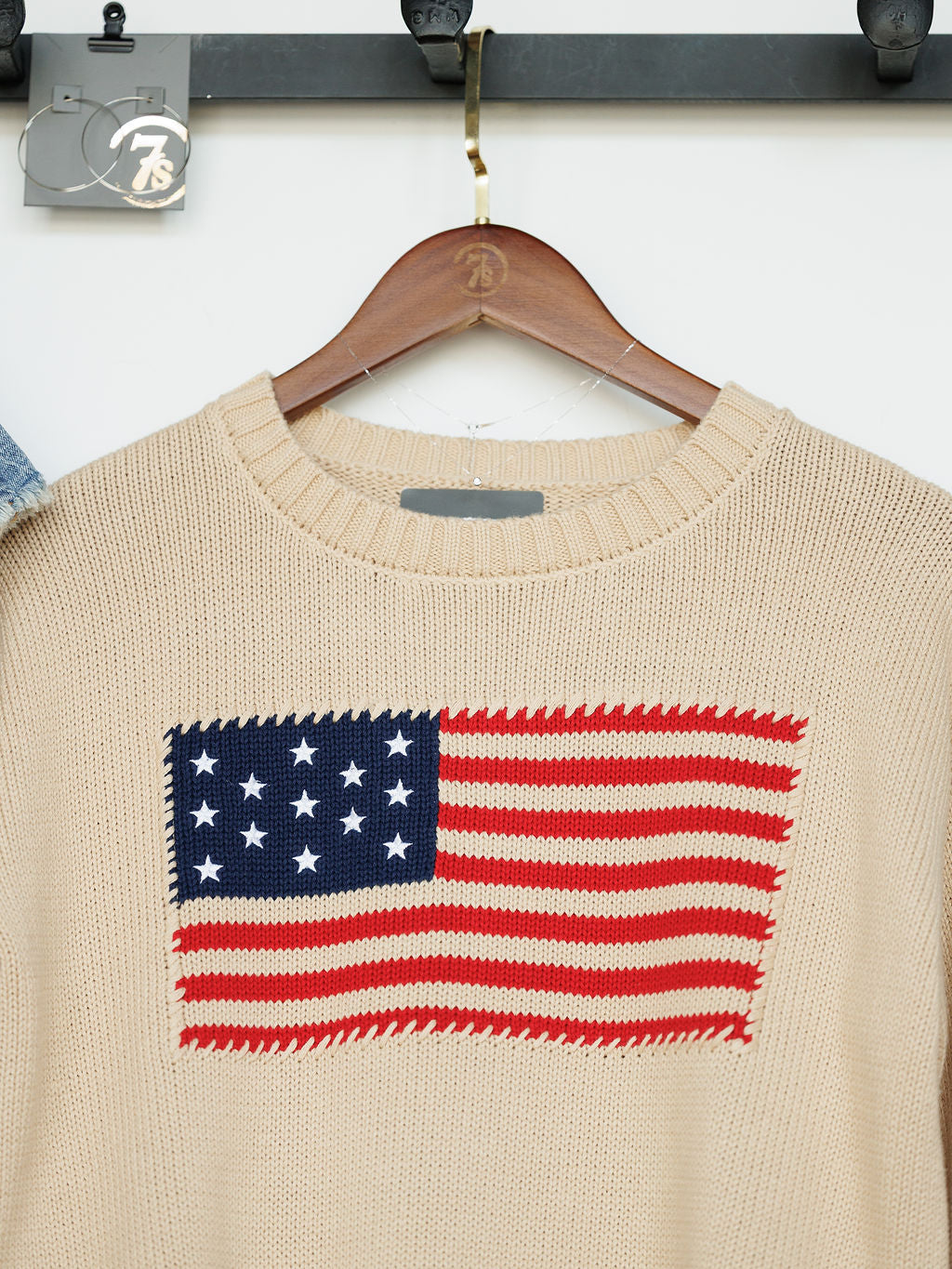 The Original {Exclusive} by Savannah Sevens western life{&}style is a beige 100% cotton embroidered sweater with a large stitched American flag patch, displayed on a wooden hanger.
