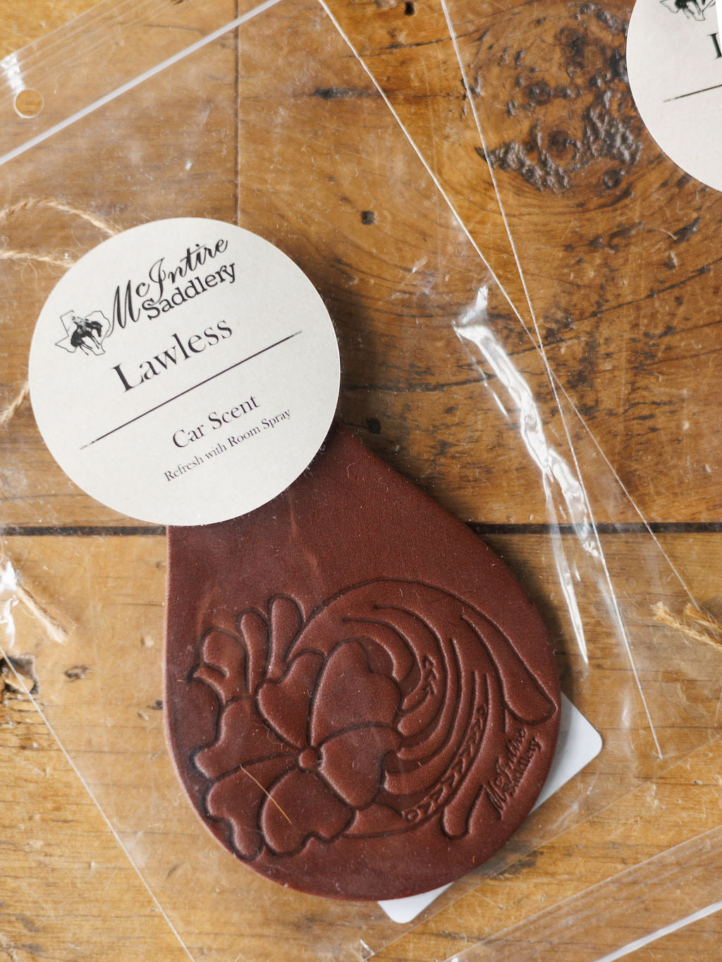 A Tooled Leather Car Freshener by Savannah Sevens western life{&}style, available in 7 scents, features a brown floral-embossed design and comes in clear packaging, displayed on a wooden surface.