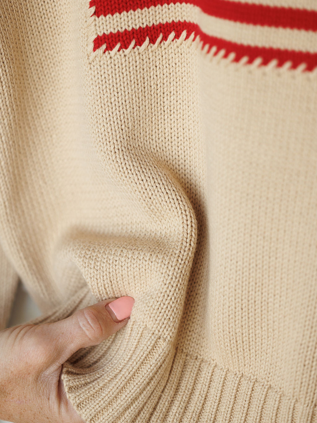 A hand with light pink nail polish pinches the hem of The Original {Exclusive} 100% cotton beige knit sweater with red stripes by Savannah Sevens western life{&}style, highlighting its texture and knitting details.
